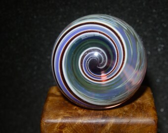 Könnte beinhalten: Eine Glaskugel mit einem wirbelnden Muster aus Weiß, Blau, Grün und Braun. Die Kugel hat eine glänzende Oberfläche und liegt auf einer Holzoberfläche.