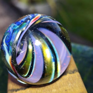 May include: A spherical glass marble featuring swirling patterns of deep blue, vibrant green, metallic gold, and soft lavender. The surface is smooth and reflective, with light catching the internal layers of color to create a multi-dimensional effect.