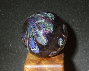 May include: A dark purple and green glass marble with a swirling pattern. The marble is sitting on a wooden base.