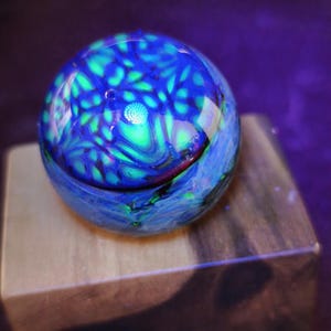 Könnte beinhalten: Ein lebendiger, kugelförmiger Glasbriefbeschwerer. Der Globus weist ein wirbelndes Muster aus Blau- und Grüntönen mit einem Hauch von Rot auf. Er ruht auf einem kleinen Holzblock und erzeugt einen auffälligen Kontrast von Texturen und Farben. Der Gesamteffekt ist eine fesselnde Darstellung von Licht und Farbe.