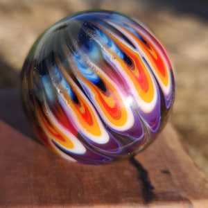 May include: A spherical glass marble featuring a swirling pattern of vibrant colors including red, orange, yellow, blue, and purple against a dark, translucent base. The surface is smooth and reflective, showcasing intricate, flame-like designs.