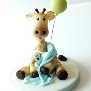 May include: A fondant giraffe cake topper holding a small stuffed animal and a green balloon. The giraffe is wearing a blue blanket.