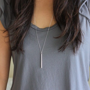 May include: A silver bar pendant necklace. The necklace features a delicate chain and a vertical bar pendant. The pendant is a simple, polished silver. The necklace is shown against a gray top.
