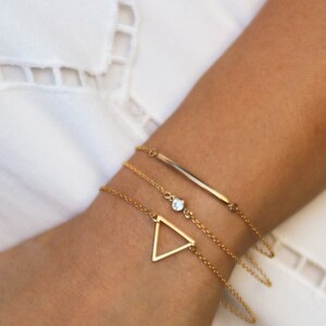 May include: A set of three gold bracelets with a delicate chain design. The bracelets feature a small triangle charm, a single clear stone, and a thin bar charm.