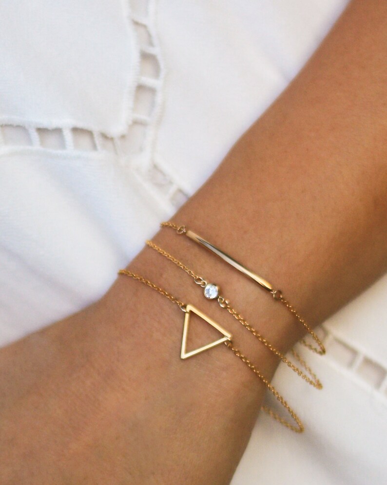 May include: A set of three gold bracelets with a delicate chain design. The bracelets feature a small triangle charm, a single clear stone, and a thin bar charm.
