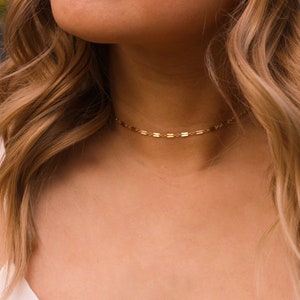 May include: A delicate gold chain choker necklace with a simple, minimalist design. The chain is made of small, flat, rectangular links.