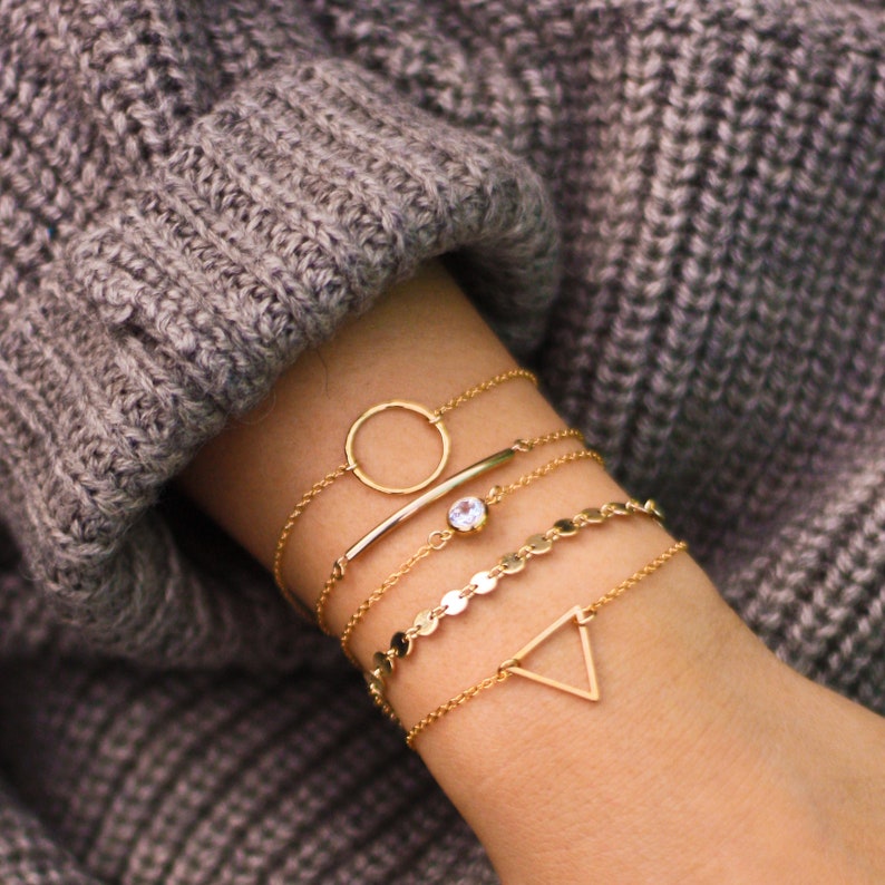 May include: A set of five gold bracelets, including a circle, a bar, a chain with small discs, a chain with a single stone, and a triangle. The bracelets are stacked on a person's wrist.