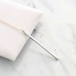 May include: A silver bar pendant necklace. The pendant is a simple, rectangular bar suspended from a delicate chain. The necklace is displayed on a white, textured surface against a marble background. The pendant is approximately 2 inches long.