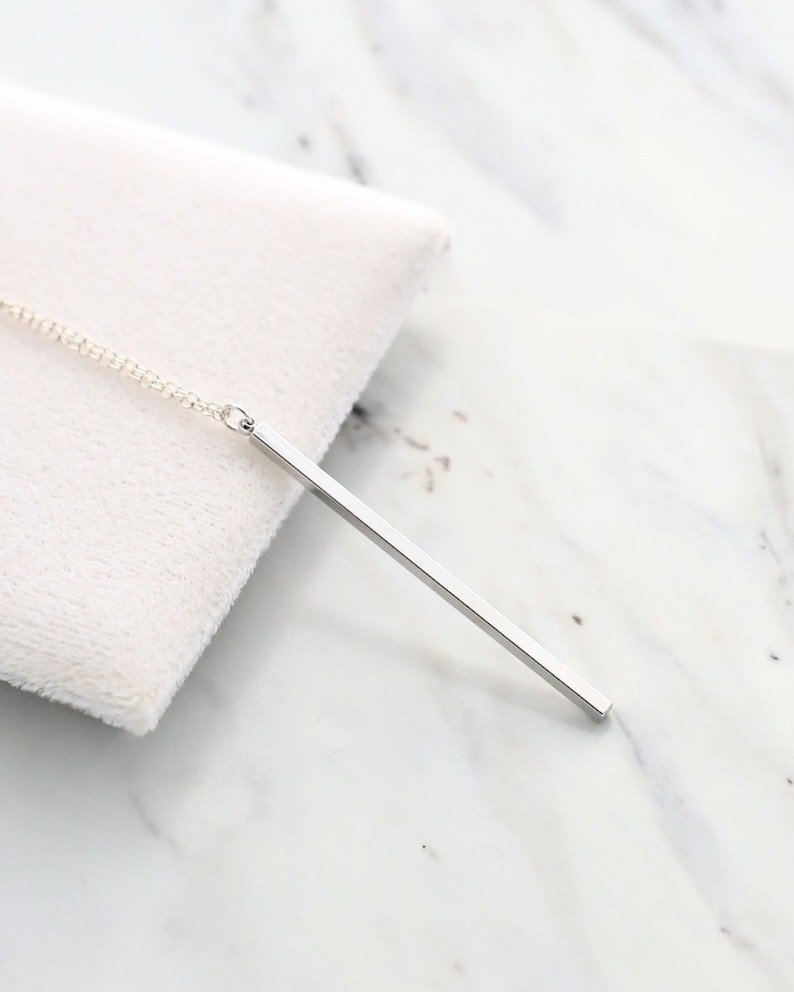 May include: A silver bar pendant necklace. The pendant is a simple, rectangular bar suspended from a delicate chain. The necklace is displayed on a white, textured surface against a marble background. The pendant is approximately 2 inches long.