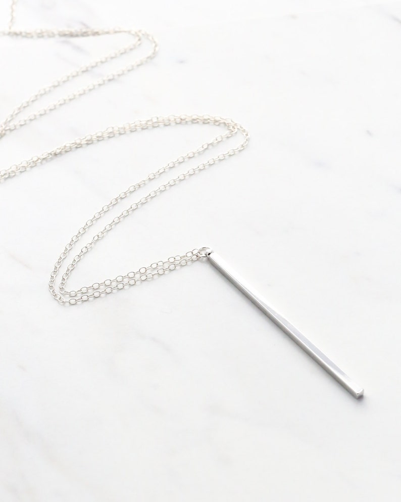 May include: A silver-colored necklace with a long, rectangular pendant. The chain is delicate and the pendant is a simple, polished bar. The necklace is displayed on a white marble surface.