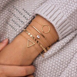 May include: A set of gold bracelets with different designs, including a circle, a bar, a triangle, and a chain with small round charms. The bracelets are stacked on a person's wrist.