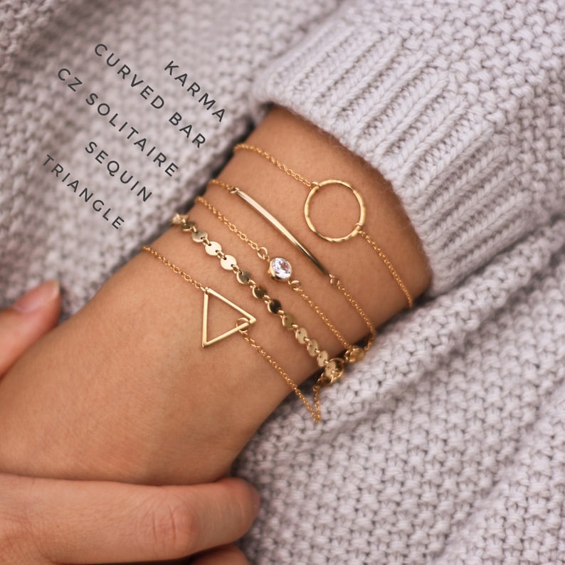 May include: A set of gold bracelets with different designs, including a circle, a bar, a triangle, and a chain with small round charms. The bracelets are stacked on a person's wrist.
