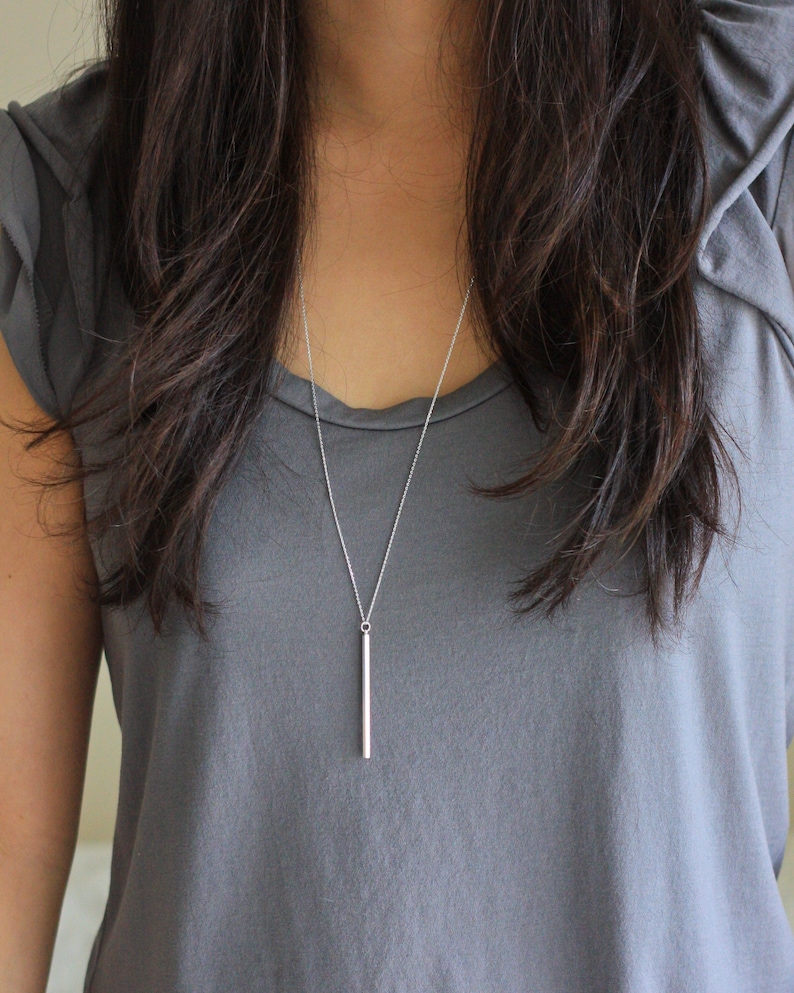 May include: A silver bar pendant necklace. The necklace features a delicate chain and a vertical bar pendant. The pendant is a simple, polished silver. The necklace is shown against a gray top.