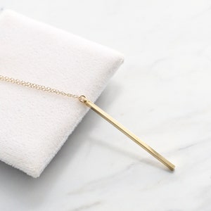 May include: A gold-colored bar pendant necklace. The pendant is a long, rectangular shape suspended from a delicate chain. The necklace is displayed on a white, textured surface, set against a marble background.