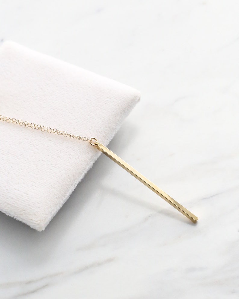 May include: A gold-colored bar pendant necklace. The pendant is a long, rectangular shape suspended from a delicate chain. The necklace is displayed on a white, textured surface, set against a marble background.