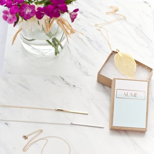 May include: A collection of necklaces on a white marble surface. The necklaces include a gold leaf pendant in a box, a gold bar pendant, a silver bar pendant, and a gold circle pendant. A vase of pink flowers is in the background.