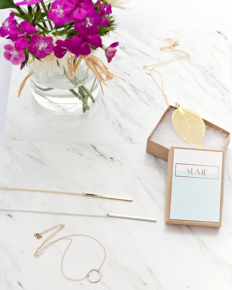 May include: A collection of necklaces on a white marble surface. The necklaces include a gold leaf pendant in a box, a gold bar pendant, a silver bar pendant, and a gold circle pendant. A vase of pink flowers is in the background.