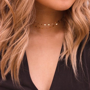 May include: A gold chain necklace with two strands, worn as a choker. The necklace is made of small, flat, rectangular links. The model is wearing a black top.