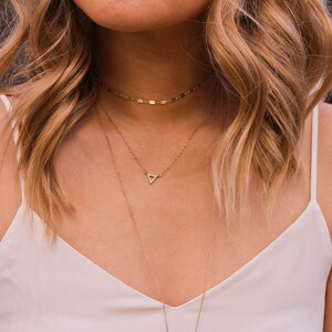 May include: A woman wearing a white tank top with two gold necklaces. The top necklace is a delicate chain with a small triangle pendant. The bottom necklace is a thicker chain.