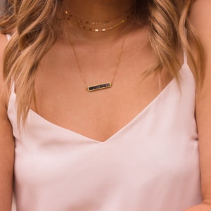May include: A gold necklace with a black bar pendant. The necklace is layered with two other gold chains.