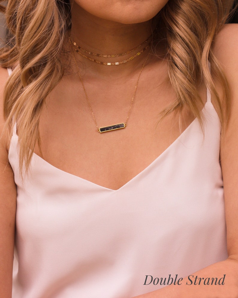 May include: A gold necklace with a black bar pendant. The necklace is layered with two other gold chains.