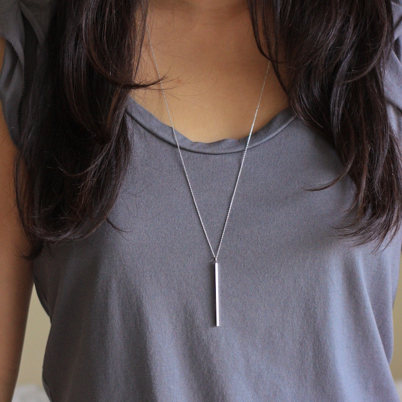 May include: A silver bar pendant necklace. The necklace features a long, rectangular pendant suspended from a delicate chain. The pendant is a simple, polished silver bar, and the chain is a fine, textured design.