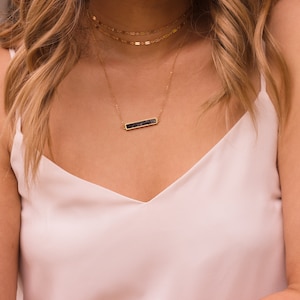 May include: A gold chain necklace with a black bar pendant. The necklace is layered with two other gold chains.
