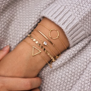 May include: A set of six gold bracelets with different designs, including a circle, a triangle, a bar, and a chain with small round charms. The bracelets are stacked on a person's wrist.