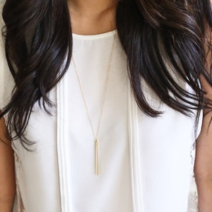 May include: A gold-colored bar pendant necklace. The necklace hangs from a delicate chain, with the bar pendant suspended at the center. The necklace is worn over a white blouse.