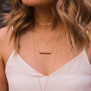 May include: A woman wearing a white tank top with a layered gold necklace set. The necklace set includes a delicate chain with a small triangle pendant, a thicker chain with a bar pendant, and a chain with a small chain link choker.