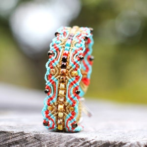 Puede incluir: Un brazalete de macramé colorido con hilos turquesa, rojo y marrón, acentuado con pequeñas cuentas.