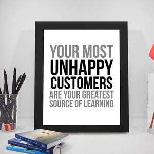 Op de afbeelding: Een zwarte fotolijst met de tekst "YOUR MOST UNHAPPY CUSTOMERS ARE YOUR GREATEST SOURCE OF LEARNING." Het frame staat op een wit oppervlak met boeken, potloden en een rode bloem in een vaas.