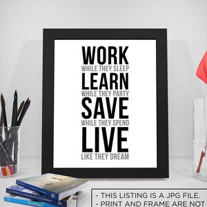 May include: Black and white typography print with the text "Work while they sleep, Learn while they party, Save while they spend, Live like they dream". The print is framed in a black frame and is displayed on a white wall.