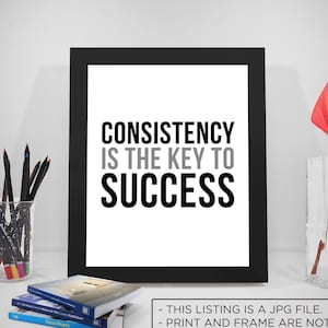 Puede incluir: Una impresión enmarcada en negro con el texto "CONSISTENCY IS THE KEY TO SUCCESS" en letras blancas sobre un fondo blanco. La impresión está sobre una mesa con un jarrón de flores rojas y un tarro de vidrio de lápices de colores.