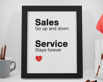 May include: Black framed print with white background and black text that reads "Sales Go up and down Service Stays forever" with a red heart symbol. The print is sitting on a white surface with pencils and a vase with a red flower.