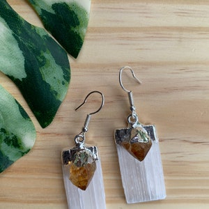 May include: A pair of silver earrings with a white selenite crystal and a golden citrine crystal. The earrings are hanging from silver hooks.