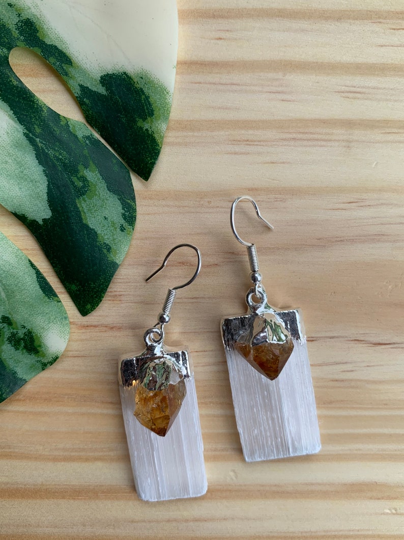 May include: A pair of silver earrings with a white selenite crystal and a golden citrine crystal. The earrings are hanging from silver hooks.