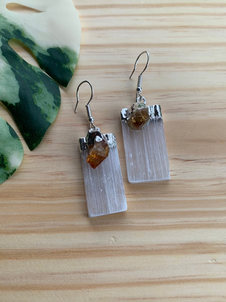 May include: A pair of silver earrings with white selenite crystal and a small amber stone. The earrings are rectangular in shape and have a silver hook.