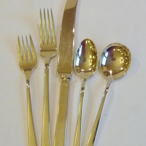 May include: A set of gold-plated silverware, including a knife, fork, and two spoons. The silverware is arranged on a white surface.