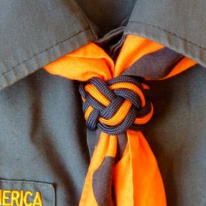 May include: A close-up of a scout neckerchief tied in a knot. The neckerchief is orange and black with a dark blue knot.