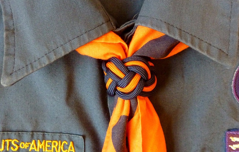 May include: A close-up of a scout neckerchief tied in a knot. The neckerchief is orange and black with a dark blue knot.