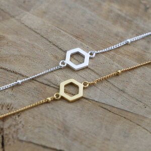 May include: Two delicate bracelets with hexagon-shaped charms. One bracelet is silver with a silver charm, and the other is gold with a gold charm.