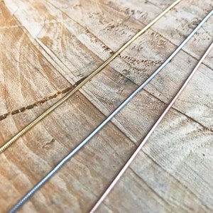 May include: Three snake chains in gold, silver, and rose gold. The chains are laid out on a wooden surface.