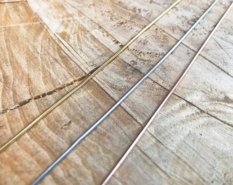 May include: Three snake chains in gold, silver, and rose gold. The chains are laid out on a wooden surface.