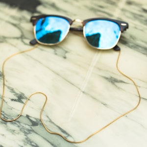 May include: A pair of blue mirrored sunglasses with a gold chain attached to the arms. The chain is lying on a marble table.