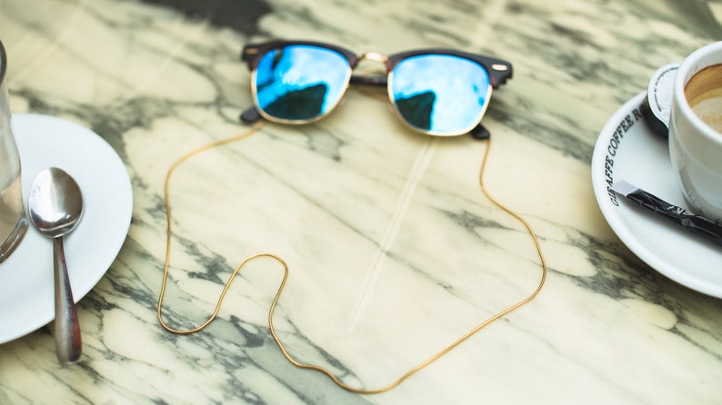 May include: A pair of blue mirrored sunglasses with a gold chain attached to the arms. The chain is lying on a marble table.