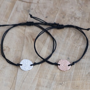 Op de afbeelding: Twee zwarte koordarmbanden met een zilveren en een roségouden ronde hanger. De hangers hebben een gehamerde textuur.