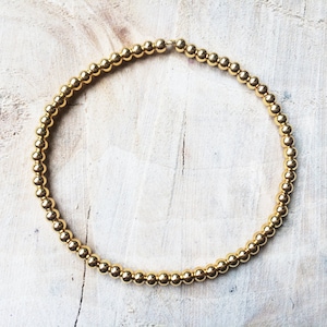 Könnte beinhalten: Ein goldfarbenes Perlenarmband, bestehend aus kleinen, gleichmäßig verteilten, runden Perlen. Das Armband ist kreisförmig und liegt auf einer hellen, strukturierten Oberfläche. Die Perlen haben ein glänzendes, metallisches Finish.