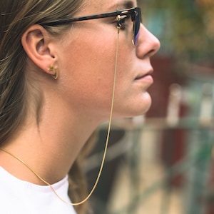 May include: A woman wearing glasses with a gold chain attached to the frames. The chain is hanging down her neck. She is wearing a white shirt and has her hair pulled back.