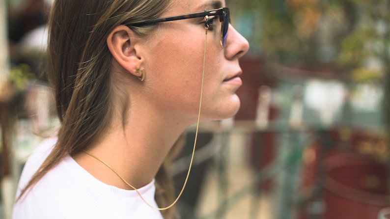 May include: A woman wearing glasses with a gold chain attached to the frames. The chain is hanging down her neck. She is wearing a white shirt and has her hair pulled back.
